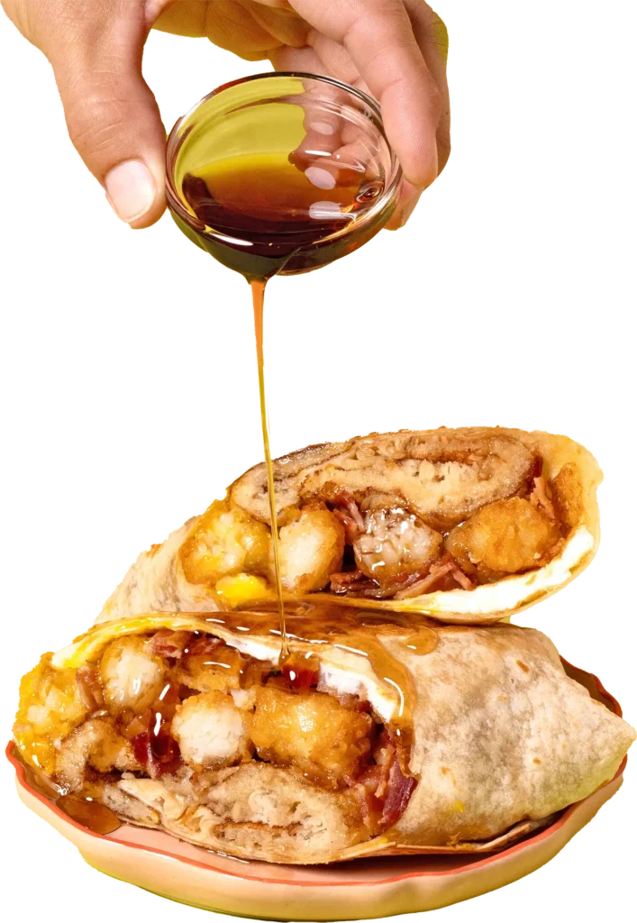 A close-up of amber syrup being poured from a small glass bowl onto a stacked breakfast burrito filled with tater tots and bacon.