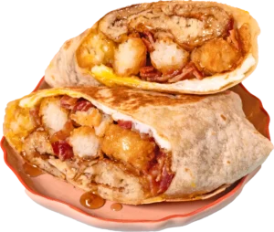 A stacked breakfast burrito on a peach-colored plate, filled with crispy chicken, tater tots, and bacon with maple syrup droplets.